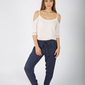 May include: A woman is wearing a white off-the-shoulder top with lace trim and dark blue jogger pants. She is wearing white sneakers.