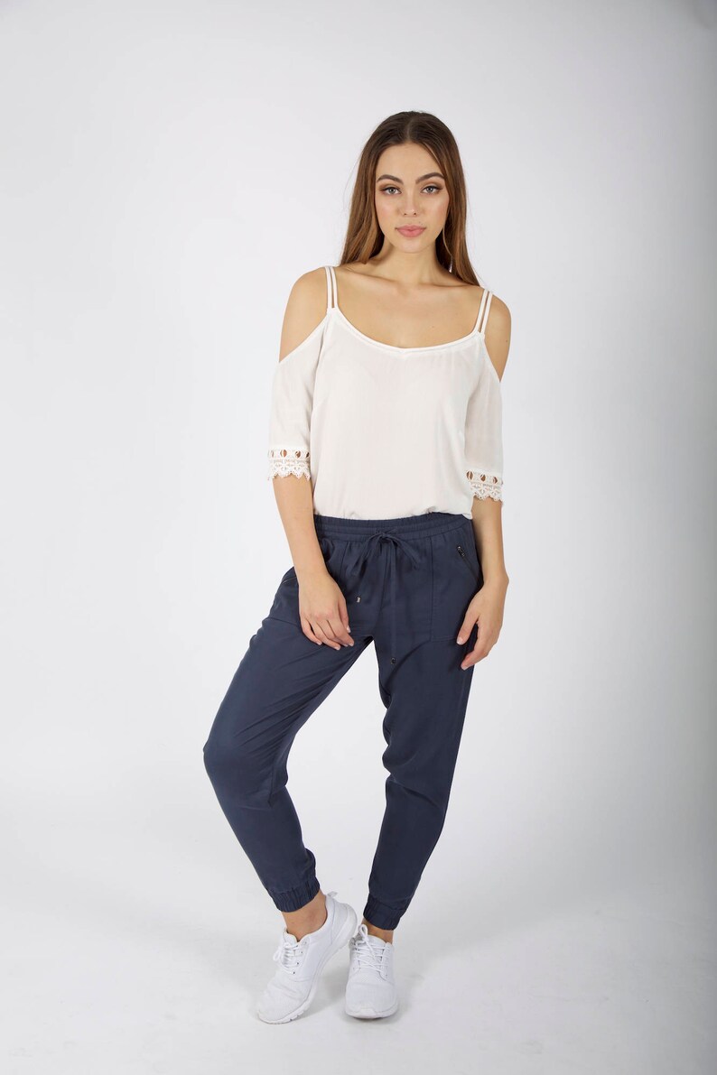 May include: A woman is wearing a white off-the-shoulder top with lace trim and dark blue jogger pants. She is wearing white sneakers.