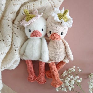 May include: Two crocheted duck plushies with flower crowns. One duck is white with a pink flower crown and the other is light pink with a green flower crown. Both ducks have orange legs.