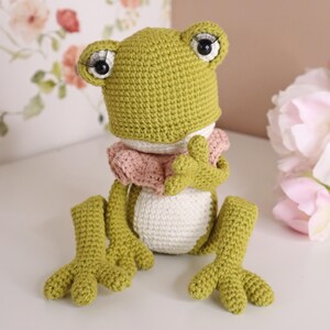 May include: A handmade crochet frog doll in shades of green, white, and pink. The frog has large eyes with eyelashes, a pink collar, and webbed feet. The frog is sitting on a white surface.