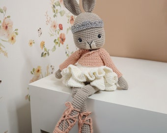 May include: A crocheted bunny wearing a pink and white ballerina costume. The bunny has long legs and is sitting on a white surface.