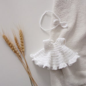 May include: White crocheted baby dress with a ruffled hem, lying on a white fuzzy blanket with wheat stalks in the background.