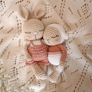 May include: A crocheted bunny and lamb doll laying together on a white crocheted blanket. The bunny is wearing a pink and white outfit and the lamb is wearing a pink and white outfit.