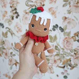 Peut inclure: Une poupée en crochet en forme de bonhomme en pain d'épices avec du glaçage blanc, du houx rouge et vert et un col à volants rouge. La poupée tient un bouton dans sa main.