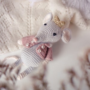 Peut inclure: Une souris en crochet gris portant une veste rose et une couronne dorée. La souris est assise sur une surface blanche.