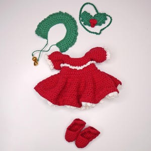 May include: A handmade crochet doll outfit in red with white trim, including a dress, shoes, a collar with bells, and a holly-shaped headpiece. The outfit is designed for a doll and is displayed on a white surface.