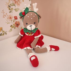 Puede incluir: Muñeca de reno de ganchillo hecha a mano con un vestido rojo, ribete blanco y cuello verde. La muñeca tiene un cuerpo marrón, cuernos blancos y una diadema verde con un lazo rojo. La muñeca lleva zapatos rojos con ribete blanco.
