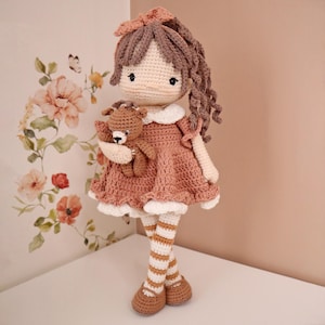 May include: A handmade crochet doll with brown yarn hair, wearing a dusty rose dress with a white collar and trim. The doll has striped white and brown legs, brown shoes, and holds a small brown crochet teddy bear in its arms.