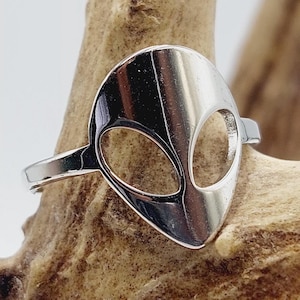 May include: A silver ring with an alien face design. The ring is on a wooden background.