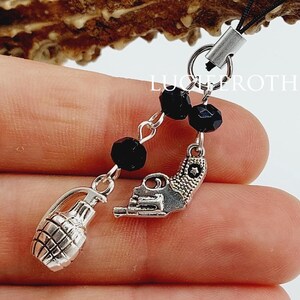 May include: A black beaded charm with silver-toned charms of a grenade and a pistol. The charm is attached to a black cord. The background includes a brown antler.