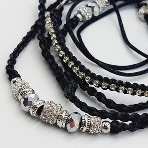 Puede incluir: Un conjunto de cinco pulseras de cordón negro con cuentas plateadas y pedrería. Las pulseras están superpuestas y atadas juntas con un nudo.