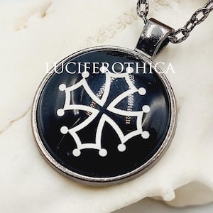 May include: A round pendant necklace featuring a black circular insert with a white Occitan cross symbol. The pendant is held by a dark metallic chain. The text LUCIFEROTHICA is printed in white above the cross symbol on the black background.