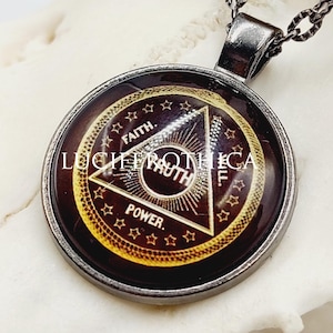 May include: A round pendant necklace featuring a dark background with a gold-toned triangle containing the words FAITH, TRUTH, and POWER. The pendant is set in a dark metal frame and hangs from a matching chain, displaying the text LECHEROTHECA.