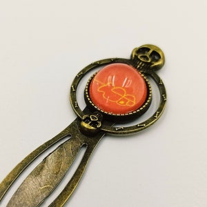 May include: A bronze bookmark with a skull design and a round, orange glass cabochon with a white design.