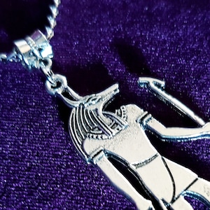 May include: Silver pendant necklace featuring an Egyptian god Anubis with a jackal head, holding a staff. The pendant is on a purple velvet background.