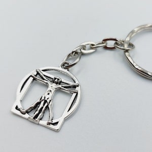 Op de afbeelding: Een zilverkleurige metalen sleutelhanger met een ronde bedel met een uitgesneden ontwerp van de Vitruviusman. De bedel is met een kort metalen kettinkje bevestigd aan een standaard sleutelring. Het ontwerp toont een menselijke figuur.