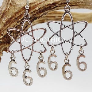 Może przedstawiać: Srebrne kolczyki z motywem atomu i wiszącymi zawieszkami z cyfrą sześć. Kolczyki zawieszone są na haczykach. Prezentowane są na jasnym tle i brązowym porożu.