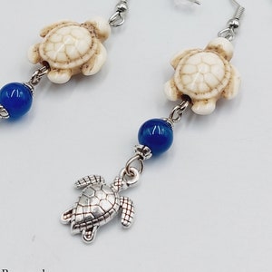 Op de afbeelding: Een paar oorhangers met een witte schildpadbedel, een blauwe kraal en een zilveren schildpadbedel. De oorhangers zijn gemaakt met zilveren haakjes.