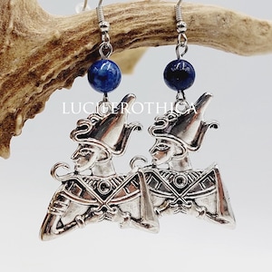 May include: A pair of silver-toned earrings featuring a stylized figure with a headdress and intricate details. Each earring has a dark blue bead at the top. The background includes a deer antler and the text "LUCIFEROTHICA".