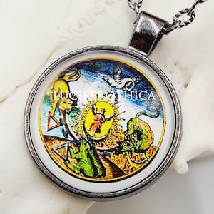 May include: A round pendant necklace featuring a dark metal frame and a chain. The pendant displays a colorful circular illustration with a central winged figure, a green frog, a yellow lion, and geometric triangles against a blue background.
