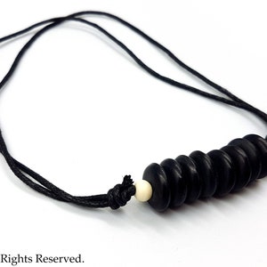 Op de afbeelding: Een zwarte koordketting met een hanger gemaakt van gestapelde zwarte kralen en een enkele witte kraal.