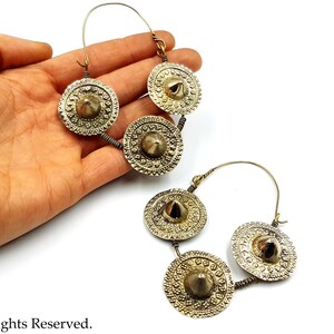 May include: A pair of gold hoop earrings with three decorative round charms on each earring. The charms have a raised center and intricate designs.