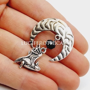 Puede incluir: Un colgante plateado con una luna creciente con superficie texturizada, un pequeño murciélago y una cuenta negra. El colgante se sostiene en una mano sobre un fondo blanco. El texto "LUCIFEROTHICA" es visible.