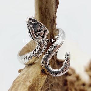 Könnte beinhalten: Ein silberfarbener Metallring in Form einer gewundenen Schlange mit strukturierten Schuppen und einem definierten Kopf. Der Ring trägt den Schriftzug LUCIFEROTHICA in der Mitte. Der Schlangenkörper windet sich zu einem offenen Ringdesign.