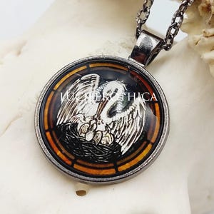 May include: A round pendant necklace featuring a silver-toned metal frame and a glass cabochon. The image inside depicts a white pelican feeding its young in a nest, surrounded by an amber and black border. The text LUCIFER ETHICA is visible.