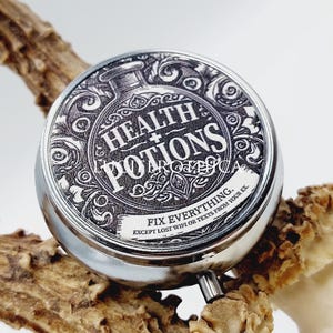 Op de afbeelding: Een ronde, zilverkleurige pillendoos met een zwart-witte afbeelding met de tekst "Health Potions". Het ontwerp bevat een illustratie van een drankfles en de tekst "Fix Everything". De doos ligt op een bruin, getextureerd oppervlak.
