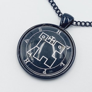 May include: A black metal necklace with a round pendant featuring a white sigil and the letters 'ASHAPH' around the edge.