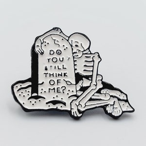 May include: Black and white enamel pin featuring a skeleton hugging a tombstone with the text "Do you still think of me?"
