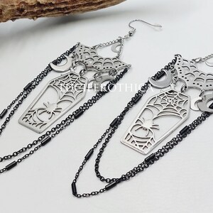May include: A pair of silver-toned dangle earrings featuring a coffin-shaped charm with a spider and web cutout design. The earrings include crescent moon accents, a web-patterned top piece, and long black chains hanging from the sides.