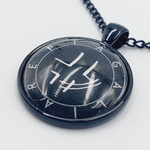 Pode incluir: Um colar preto com um pingente redondo que apresenta um selo branco e as letras "RE-P-A-G-A-A" ao redor da borda.