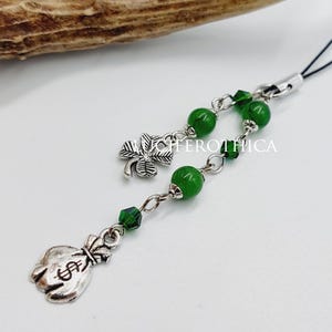 Op de afbeelding: Een zilveren bedel met een klavertjevier en een geldbuidel hanger, verbonden door een ketting van groene kralen. De bedel is bevestigd aan een zwarte lus. Een hertengewei staat op de achtergrond.