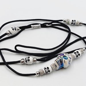 Puede incluir: Un collar de cordón negro con cuentas de calavera blancas y detalles plateados. La cuenta central de la calavera está pintada con detalles coloridos. Cierre de cierre.