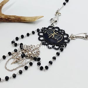 Op de afbeelding: Een gotische ketting met een zwarte en zilveren vleermuis hanger, zwarte kralenkettingen en handvormige accenten. De ketting heeft een donkere esthetiek, met een ovale hanger en ingewikkelde details.
