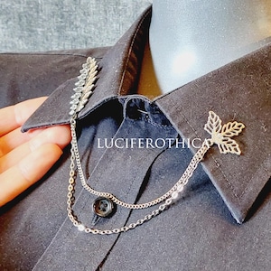 Op de afbeelding: Zilverkleurige kraagketting met blad- en bloemaccenten, bevestigd aan een zwart overhemd met knopen. De ketting verbindt twee decoratieve elementen op de kraag. De tekst "LUCIFEROTHICA" is zichtbaar.