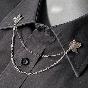 May include: A silver-coloured collar chain with leaf-shaped details. The chain links two leaf-shaped pins, designed to be attached to a shirt collar. The shirt is black.