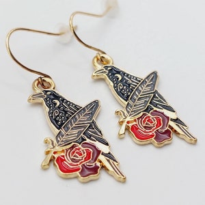 Puede incluir: Pendientes de oro con detalles de cuervo negro y rosa roja. El cuervo sostiene una pluma en su pico y la rosa tiene un tallo verde.