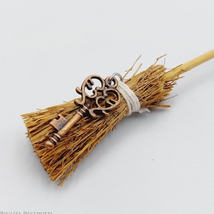 May include: A miniature broom with a brown wooden handle and a straw broom head. A decorative antique-style key is attached to the broom head.