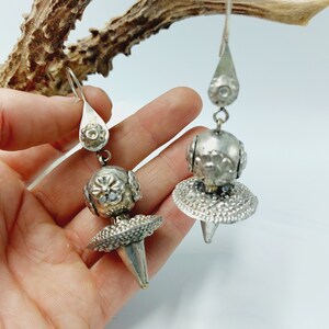 May include: A pair of silver dangle earrings with a round, textured ball and a pointed, textured cone. The earrings are hanging from a silver hook.