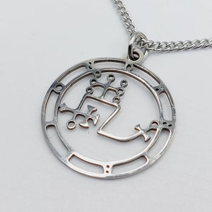 May include: Silver necklace with a circular pendant featuring a detailed, intricate design. The pendant is made of metal and has a cutout design.