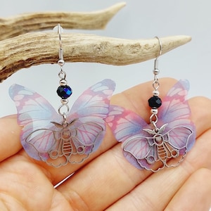 Op de afbeelding: Een paar zilveren vlinder oorringen met een delicaat, doorschijnend roze en blauw ontwerp. Elke vlinder heeft een zwarte kristallen kraal die eraan hangt.