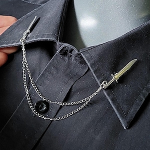 May include: Silver-toned collar chain with a small knife pendant. The chain is draped across the collar of a black button-down shirt. The knife pendant has a detailed blade and handle.