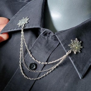 Op de afbeelding: Zilverkleurige kraagketting met zonvormige accenten. De ketting heeft drie strengen en is bevestigd aan de kraag van een zwart overhemd met knopen. Het zonvormige ontwerp heeft een centraal, cirkelvormig element.