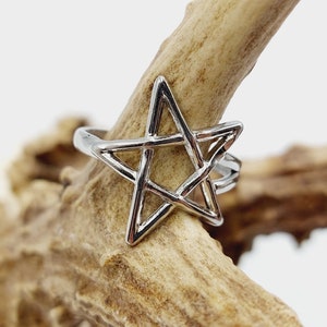May include: A silver-colored ring featuring a five-pointed star design. The ring is set against a blurred background of a light brown, textured surface. The star is the focal point, with its geometric shape clearly defined.