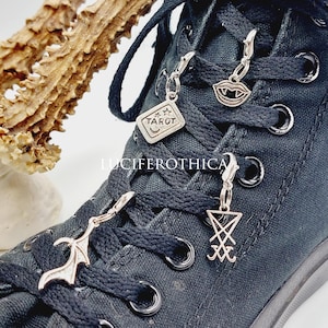 Op de afbeelding: Close-up van een zwarte canvas schoen versierd met zilveren bedels. Bedels zijn onder meer een tarotkaart, vampierlippen, een vleermuisvleugel en een sigil. De schoenveters zijn zwart.