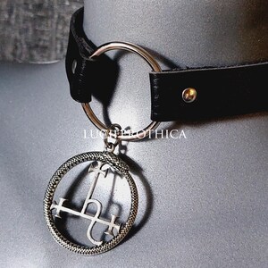 Könnte beinhalten: Ein schwarzes Lederhalsband mit einem silberfarbenen Metallring und einem kreisförmigen Anhänger, der eine sich in den Schwanz beißende Schlange zeigt, die ein Luzifer-Siegel umschließt. Der Text LUGHLOTHICA ist in der Mitte des Designs zu sehen.
