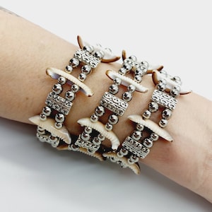 Op de afbeelding: Een drievoudig armband met witte schelpbedels en zilveren kralen. De bedels zijn halfrond en hebben een getextureerd ontwerp. De kralen zijn rond en hebben een glanzende afwerking.